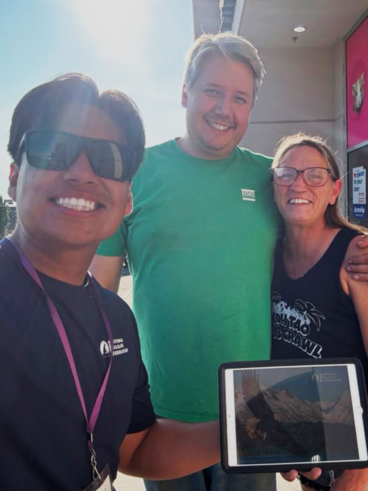 Smiling person wearing lanyard and NWF t-shirt and holding iPad with NWF image on the screen, standing next to two other smiling people outside of retail store
