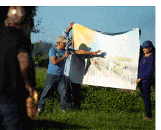 Two people holding up banner depicting agricultural worker working under a large sun while a speaker holding a microphone stands in front of the banner and points to it