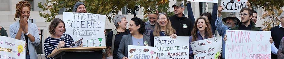People holding signs at the Voices for Conservation Rally while Tara Losoff speaks into a microphone at the lectern